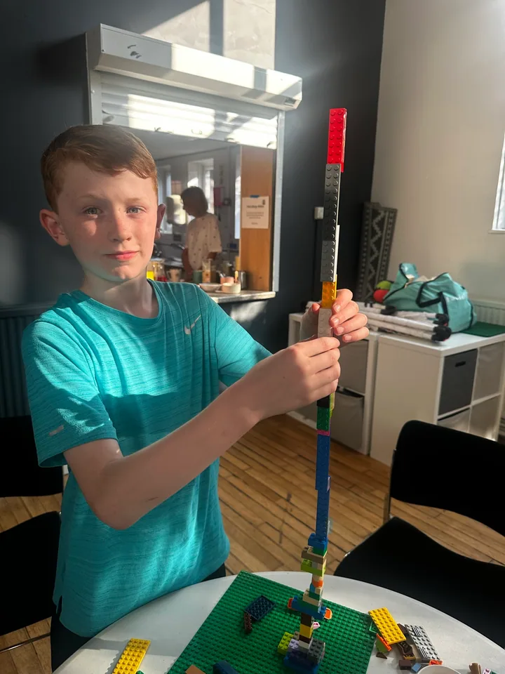 A young builder holding a tall brick tower beside a green baseplate.