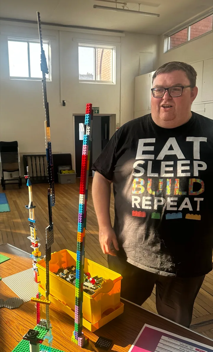 A builder in an Eat Sleep Build Repeat T-shirt beside a tall multicoloured tower.